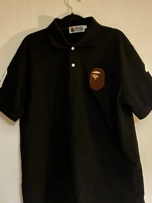 Bape Polo svart - Säljer en svart t-shirt från BAPE med klassisk ap-logga på bröstet och vita siffror på ärmarna. T-shirten har krage, knappar framtill och är kortärmad. Perfekt för dig som gillar streetwear och vill sticka ut.