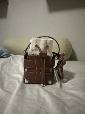 Brun bucket bag med canvasinsats - Cool brun bucket bag i läder med vita sömmar och en beige canvaspåse inuti. Väskan har både handtag och lång axelrem, samt snygga detaljer med snörning framtill. Perfekt för dig som gillar trendiga accessoarer med unik design.