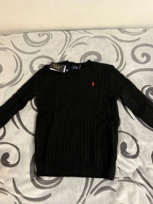 Svart kabelstickad tröja Polo Ralph Lauren - Kabelstickad svart tröja från Polo Ralph Lauren med klassisk röd logga broderad på bröstet. Tröjan är i 100% bomull, har rund halsringning och långa ärmar. Perfekt för dig som gillar stilrena och tidlösa plagg.