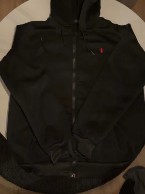 Svart hoodie från Polo Ralph Lauren - Svart hoodie med dragkedja från Polo Ralph Lauren. Klassisk design med huva och dragsnören samt röd broderad logga på bröstet. Mjuk bomullskänsla och ribbade muddar vid ärmslut och nederkant. Perfekt för en avslappnad och stilren look.