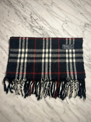 Burberrys svart-rutig halsduk  - Klassisk halsduk från Burberrys i svart, vitt och rött rutmönster. Tillverkad i mjuk ull med fransar i ändarna och ikonisk Burberrys-label. Perfekt accessoar för att lyfta din outfit med en tidlös brittisk stil.