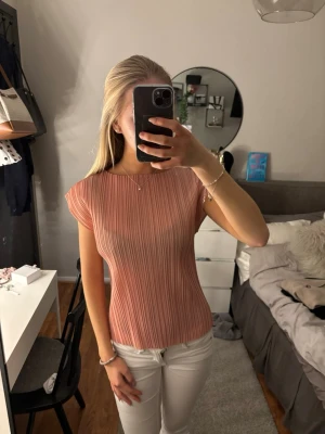 Rosa plisserad topp med kort ärm - Säljer en ljusrosa plisserad topp med korta ärmar och rund halsringning. Toppen har en slim passform och ett stilrent, ribbat mönster som ger en snygg struktur. Perfekt att matcha med jeans eller kjol för en trendig look.
