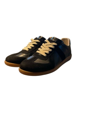 Svarta Replica sneakers med mockadetaljer - Snygga svarta Replica sneakers från Maison Margiela med kontrasterande vita skosnören och bruna gummisulor. Skorna har en mix av mocka och läder i ovandelen samt klassisk låg silhuett. Perfekta för dig som gillar stilrena och tidlösa sneakers.