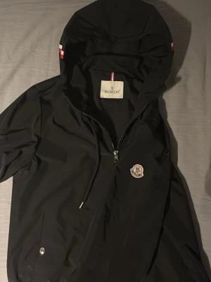 Svart vindjacka från Moncler - Snygg svart vindjacka från Moncler med huva och dragkedja framtill. Jackan har två fickor med tryckknappar, Moncler-logga på bröstet och detaljer i rött, vitt och blått på huvan. Tillverkad i ett lätt och vattenavvisande material.