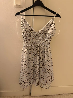 Vit prickig kort klänning med knyt - Söt vit kort klänning med svarta prickar och smock i midjan. Klänningen har tunna axelband med knyt och v-ringad hals. Perfekt för varma dagar och ger en avslappnad men trendig look.
