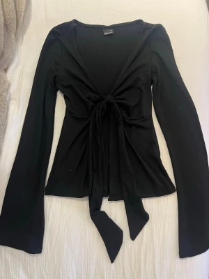Svart knytblus från Gina Tricot - Trendig svart långärmad topp från Gina Tricot med knyt framtill och v-ringad urringning. Modellen är figurnära och har långa, lätt utsvängda ärmar. 💓
