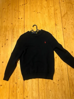 Svart stickad tröja från Ralph Lauren - Klassisk svart stickad tröja från Ralph Lauren med rund halsringning och röd broderad logga på bröstet. Tröjan har ribbade muddar vid ärmslut och nederkant. Perfekt för en stilren och enkel look.
