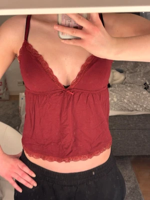 Vinrött linne från Nelly - Säljer ett oanvänt vinrött linne med tunna axelband och spetsdetaljer längs urringningen och nederkanten. Toppen har även en liten rosett framtill.