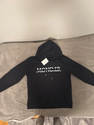 Svart Moncler hoodie med logga - Säljer en svart hoodie från Moncler med stor vit logga på bröstet och klassisk Moncler-patch på ärmen. Hoodien har huva med dragsko och en stor magficka. Tillverkad i mjukt bomullsmaterial som känns riktigt skönt mot huden. PRIS KAN DISKUTERAS