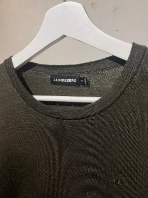  Stickad tröja från J.Lindeberg - Snygg mörkgrön långärmad tröja från J.Lindeberg med rund halsringning. Tröjan har diskret logga på bröstet och är tillverkad i ett tunt, stickat material som passar perfekt till lager-på-lager-stil.