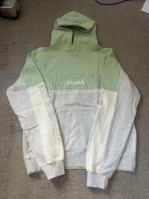 Grön och grå hoodie från H&M - Snygg hoodie från H&M i ljusgrönt och grått med texten 'WEEKEND ahead MAKING NEW MEMORIES' framtill. Hoodien har huva, ribbade muddar och paneler i olika färger. Perfekt för chill dagar och streetstyle.