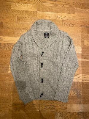 Grå cardigan  - Kabelstickad grå kofta från Ambrose Brand & Co med stora svarta knappar och bred sjalkrage. Koftan har lång ärm och ribbade muddar, perfekt för lager-på-lager och en chill look. Storlek small men passar medium 