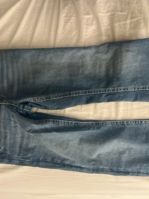 Blå bootcut jeans med broderad ficka - Säljer ett par ljusblå bootcut jeans med snygg broderad bakficka och knappdetalj. Jeansen har klassisk femficksdesign och är tillverkade i denim med lätt tvättad look. Perfekta för dig som gillar en avslappnad men ändå trendig stil.