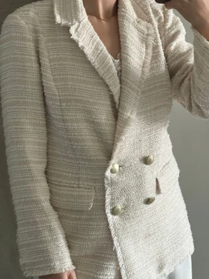 Krämvit bouclé kavaj med guldknappar - Stilren krämvit kavaj i bouclétyg med dubbelknäppning och dekorativa guldknappar. Klassisk krage, två fickor framtill och råa kanter för en trendig look. Perfekt för dig som gillar en chic och elegant stil.