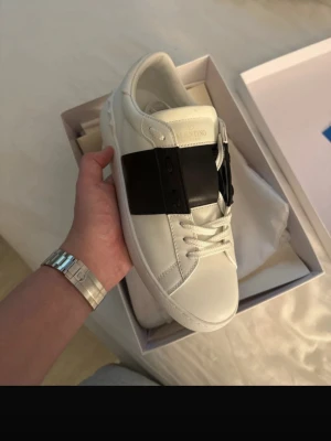 Valentino open vita sneakers - Snygga vita sneakers från Valentino Garavani med en bred svart läderdetalj över mitten. Skorna har klassisk låg profil, vita snören och diskreta nitar på sulan. Perfekta för dig som gillar stilrena och exklusiva sneakers. Kvitto finns! (Samma skor som min andra annons endast nya bilder)