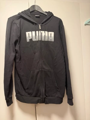 Svart zip hoodie från Puma - Säljer en svart hoodie från Puma med dragkedja framtill och stor vit logga på bröstet. Hoodien har huva med snörning och två fickor framtill. Tillverkad i mjukt och bekvämt material, perfekt för chill dagar eller när du vill ha en sportig look.