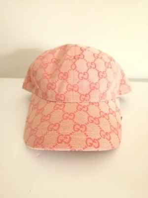 Rosa Gucci keps med GG-mönster - Säljer en snygg rosa keps Gucci med det klassiska GG-mönstret i en rosa färg över hela kepsen. Kepsen har böjd skärm och är tillverkad i ett slitstarkt tygmaterial. Perfekt för dig som vill ha en trendig och exklusiv accessoar.
