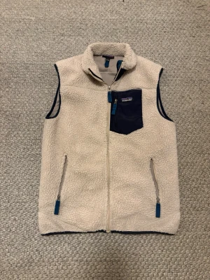 Beige fleeceväst från Patagonia - Mysig beige fleeceväst från Patagonia med marinblå detaljer. Västen har dragkedja framtill, två fickor med dragkedja och en bröstficka i marinblått tyg. Perfekt för lager-på-lager och outdoor-stil. 