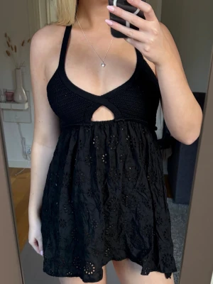 Svart kort klänning med virkad topp - Supersöt svart kort klänning med virkad topp och broderad kjol. Klänningen har en v-ringad hals, cut-out detalj framtill och breda axelband. Kjolen är dekorerad med blommiga broderier och har en luftig känsla, perfekt för sommaren och stranden🫶🏼