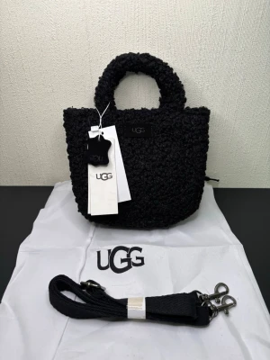 Svart teddyhandväska från UGG - Snygg svart handväska från UGG i mjukt teddy-material. Väskan har rundade handtag, avtagbar axelrem och UGG-logga framtill. Perfekt för dig som gillar trendiga accessoarer med mysig känsla. Levereras med dustbag och taggar kvar.