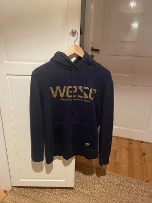 Mörkblå WESC hoodie med guldtryck - Snygg mörkblå hoodie från WESC med stort guldigt logotryck på bröstet och texten 'WeAretheSuperlativeConspiracy' under. Klassisk känguruficka framtill och justerbar huva med snören. Skriv för mer bilder. 