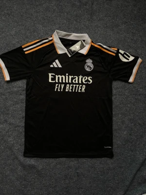 Svart Real Madrid matchtröja Adidas - Snygg svart matchtröja från Real Madrid med vita och orange detaljer. Tröjan har klubbmärke på bröstet, Adidas-logga och sponsortryck framtill. Tillverkad i ett lätt och ventilerande material, perfekt för träning eller match. Finns i S, M och L