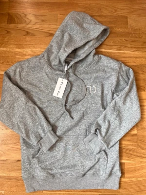 Grå hoodie  - Snygg grå hoodie med broderad logga på bröstet och klassisk känguruficka. Mjuk bomullsblandning och ribbade muddar vid ärmslut och midja. Perfekt för en chill och stilren look.