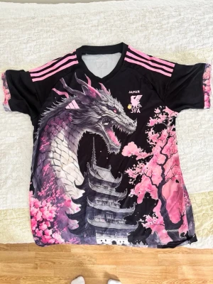 Japan JFA drake t-shirt med körsbärsblom - Unik svart t-shirt med rosa detaljer från Japan JFA. Framsidan har en stor drake, körsbärsblommor och ett japanskt tempel. Tre rosa ränder på axlarna och v-ringad hals. Äkta