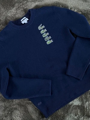 Mörkblå stickad Lacoste-tröja - Snygg mörkblå stickad tröja från Lacoste med rund halsringning och fem broderade gröna krokodiler på bröstet. Tröjan har ribbade muddar vid ärmslut och nederkant. Perfekt för dig som gillar klassisk stil med en twist.