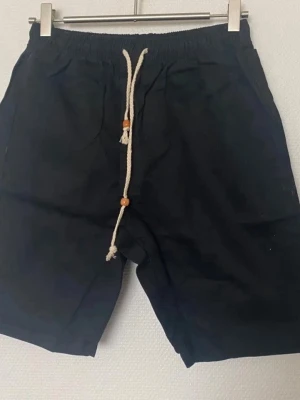 Svarta shorts med snörning - Svarta shorts med elastisk midja och vit snörning med trädetaljer. Shortsen har fickor fram och en bakficka, perfekt för en avslappnad stil. Materialet känns mjukt och passar bra till varma dagar. Helt ny , finns Stlk kvar M, L , XL och XXL 