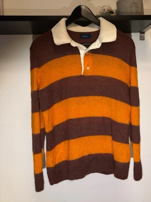 Randig stickad tröja från Gant - Stickad tröja från Gant med breda bruna och orange ränder. Klassisk vit krage och knappar vid halsen ger en retro rugbykänsla. Långärmad modell i mjukt stickat material, perfekt för höst och vinter.