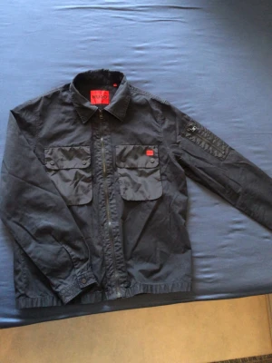 HUGO - Svart overshirt från HUGO med två bröstfickor med tryckknappar. Dragkedja framtill och klassisk krage. Röd HUGO-detalj på ena fickan. Fint skick, storlek S.