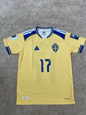 Sverige fotbollströja Gyökeres 17 Adidas - Gul Sverige fotbollströja från Adidas med blå detaljer, nummer 17 och namnet Gyökeres på ryggen. Tröjan har landslagets emblem på bröstet, korta ärmar och är tillverkad i lätt funktionsmaterial. Perfekt för match eller träning.