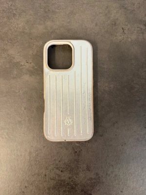 Silvrigt skal från Rimowa till iPhone 16 PRO  - Snyggt silvrigt mobilskal från Rimowa med klassisk räfflad design och diskret logga på baksidan. Skalet är gjort i tålig plast och har exakta utskärningar för knappar och kamera. Perfekt för dig som vill ha en stilren och exklusiv look på mobilen.