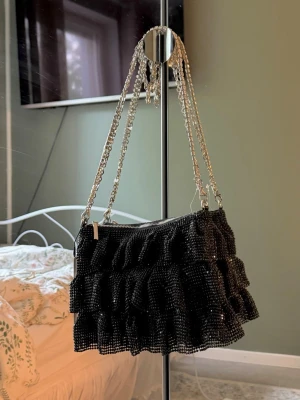 Svart axelväska med volanger och kedja - Snygg svart handväska med glittrande stenar och flera lager volanger. Väskan har en silverfärgad kedjerem och dragkedja upptill. . Materialet är tyg täckt med små glittrande stenar.