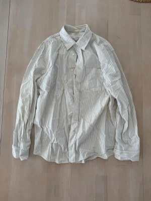Vit randig skjorta med knappar - Klassisk vit skjorta med tunna grå ränder och knäppning framtill. Skjortan har lång ärm, bröstficka och en avslappnad passform. Perfekt till jeans eller chinos för en clean och enkel look.