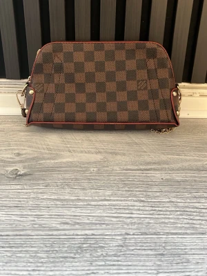 Brun L och V axelremsväska och hand väska  - Snygg handväska från L och V i klassiskt brunt damier-mönster. Väskan har guldfärgade metalldetaljer, kedjerem och röda kanter som ger en exklusiv känsla. Perfekt storlek för det viktigaste och enkel att bära över axeln. Det är en axel väska och en handväska. Kedjan går att ta bort och användas som handväska. 