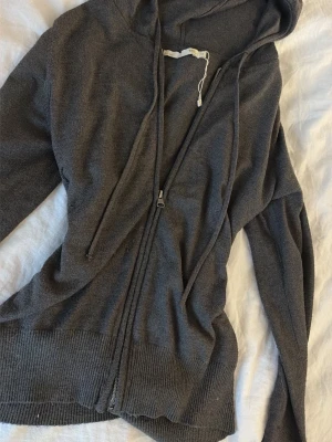 Mörkgrå hoodie med dragkedja - Mysig mörkgrå hoodie med dragkedja framtill och justerbar huva med snören. Tillverkad i mjukt stickat material och har ribbade muddar vid ärmslut och nederkant. Perfekt för en avslappnad och chill stil.