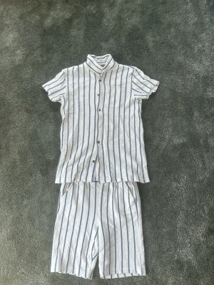 Randig pyjamas med kort ärm och shorts - Säljer en fräsch pyjamas i vitt med tunna blå ränder. Setet består av en kortärmad skjorta med knappar och bröstficka samt matchande shorts med resår i midjan. Materialet känns luftigt och mjukt, perfekt för chill dagar eller nätter.