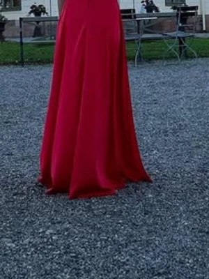 Röd långklänning med slits - Elegant röd långklänning i ett glansigt material med en hög slits på sidan. Klänningen har en figurnära passform upptill och faller snyggt längs kroppen. Perfekt för speciella tillfällen när du vill sticka ut.