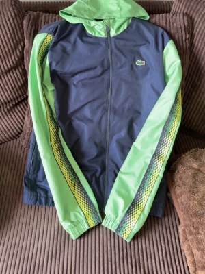Lacoste - Snygg vindjacka från Lacoste i marinblått och ljusgrönt med huva och dragkedja. Ärmarna har coola grafiska ränder i grönt, gult och svart. Jackan har den klassiska Lacoste-loggan på bröstet och är tillverkad i lätt, vindtätt material.