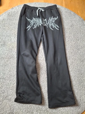 Svarta mjukisbyxor från MANNITY - Svarta mjukisbyxor från MANNITY med vitt tryck i metallstil över framsidan. Byxorna har resår i midjan med vit dragsko och raka ben. Perfekta för chill dagar eller streetwear-look. Tillverkade i mjukt material för maximal komfort.