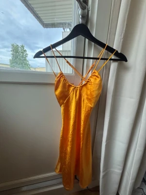 Gul omlottklänning med smala band - Snygg gul kortklänning med tunna axelband och omlottdetalj i ryggen. Klänningen har en lätt glansig yta och är perfekt för varma dagar. Materialet känns svalt och följsamt, och modellen har en feminin siluett med öppen rygg.