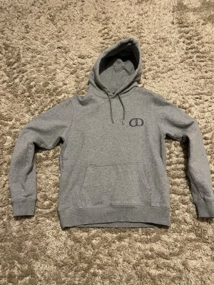 Grå hoodie med logga från dior  - Snygg grå hoodie från Oakley med klassisk känguruficka och justerbar huva. Diskret Oakley-logga broderad på bröstet. Tillverkad i mjukt bomullsmaterial som är perfekt för chill dagar. Enkel och stilren design som passar till det mesta.