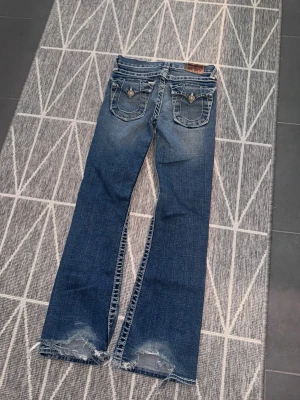 Blå bootcut jeans med slitningar flare w24 - Säljer ett par blå bootcut jeans med slitna detaljer vid bensluten och snygg tvätt. Jeansen har fickor med lock och knappar bak samt klassisk femficksdesign. Perfekta för dig som gillar en avslappnad och trendig look. w23/w24. fråga för mått. 