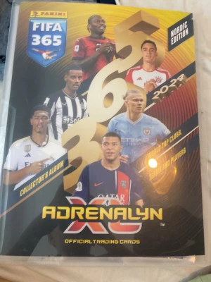 Panini FIFA 365 Adrenalyn XL samlaralbum - Säljer ett Panini FIFA 365 Adrenalyn XL samlaralbum Nordic Edition 2023/2024. Albumet innehåller officiella trading cards med fotbollsspelare från världens toppklubbar. Omslaget är svart och gult med färgbilder på spelare och text i guld och vitt.