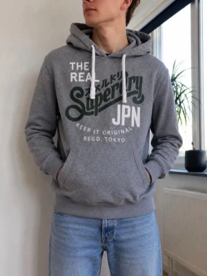 Grå Superdry hoodie med tryck - Snygg grå hoodie från Superdry med stort vitt och grönt tryck på bröstet och textdetaljer på japanska. Klassisk känguruficka framtill, vita dragsnören i huvan och ribbade muddar. Perfekt för en avslappnad streetwear-look.