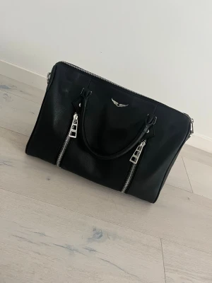 Svart handväska från Zadig & Voltaire - Cool svart handväska i läder från Zadig & Voltaire med silverfärgade detaljer och två dragkedjor framtill. Väskan har dubbla handtag och en snygg logotyp i metall. Perfekt för dig som gillar stilrena accessoarer med rockig touch. Stora modellen 