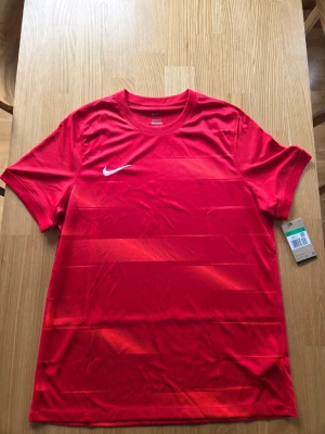 Nike röd träningströja,slim fit , storlek XL - Röd Nike Dri-FIT T-shirt för män, storlek XL. Tröjan har korta ärmar och en rund halsringning. Den är designad med Nikes Dri-FIT-teknologi för att hålla dig torr och bekväm. Tröjan har ett diskret mönster med horisontella ränder och diagonala linjer i olika nyanser av rött. Nike-logotypen är placerad på vänster bröst.