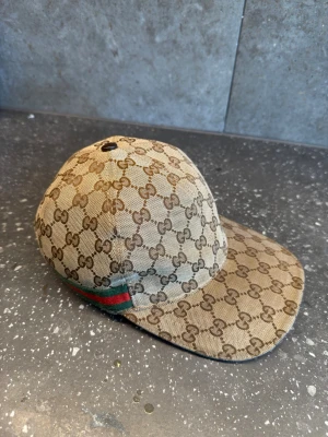Gucci beige keps med GG-mönster - Säljer en beige keps från Gucci med det klassiska GG-monogrammet över hela kepsen. Kepsen har en böjd skärm och en grön-röd rand på sidan. Tillverkad i canvasmaterial med justerbart band bak. Perfekt för dig som vill ha en lyxig och trendig accessoar.